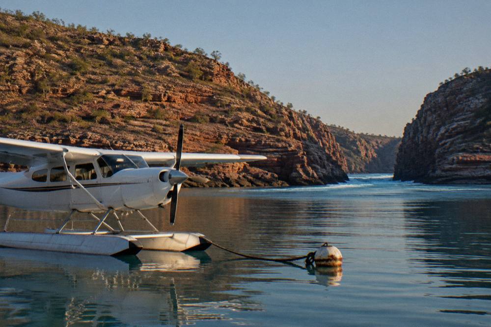 A seaplane floating on calm turquoise water beside a red rock coastline in Talbot Bay