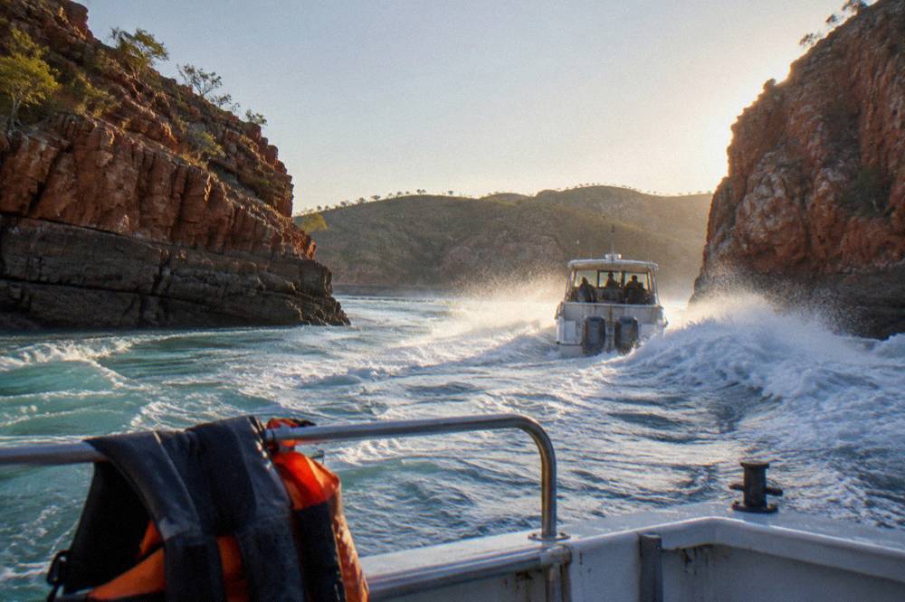 A power boat crossing white turbulent water between steep red cliffs at a tidal gap