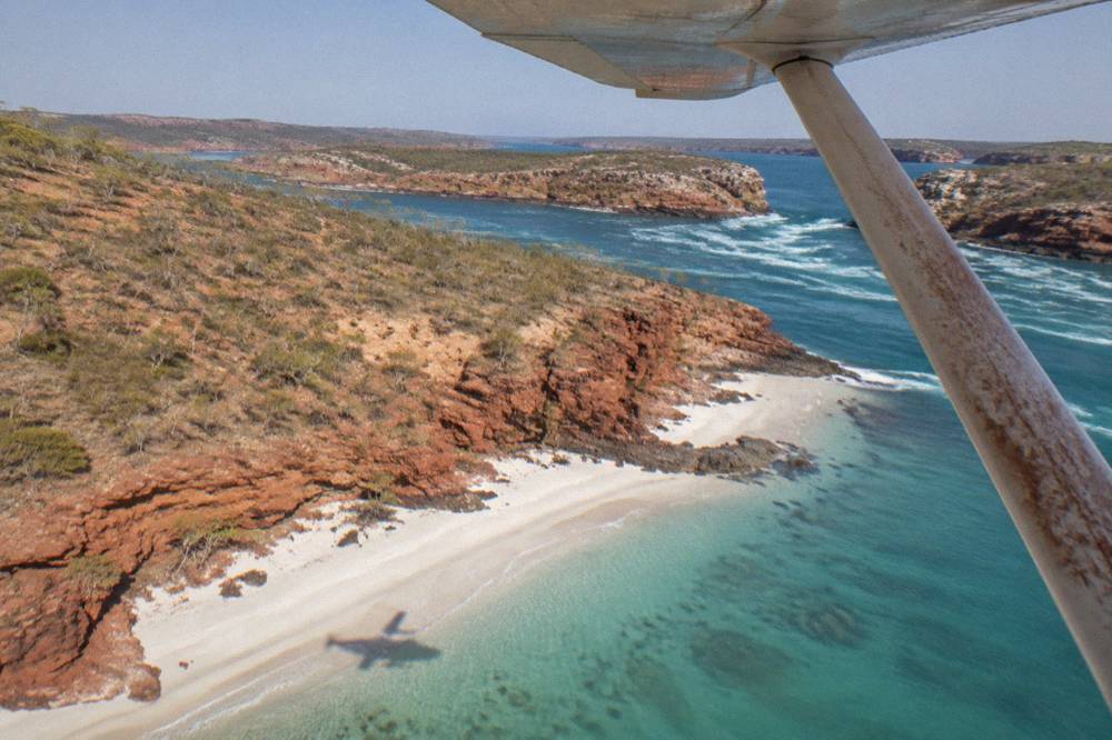 A light aircraft flying over islands and clear water along a red rock coastline