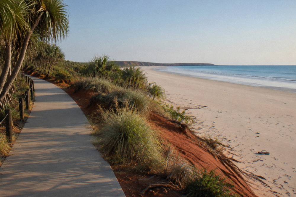 A shaded footpath beside red pindan dunes leading toward a wide empty beach at Cable Beach