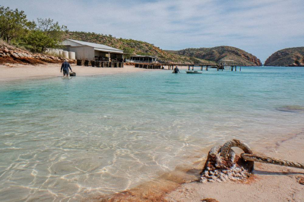 Turquoise shallows and pale sand at the edge of Cygnet Bay with pearl farm sheds in the distance