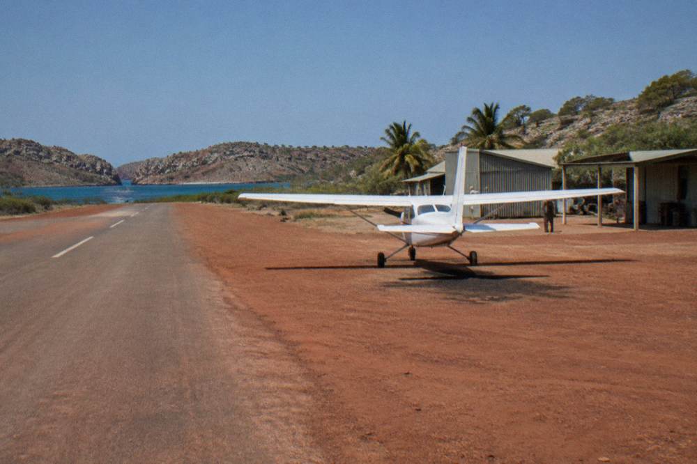 Light aircraft parked on a dirt strip beside pearl farm buildings under a wide blue sky