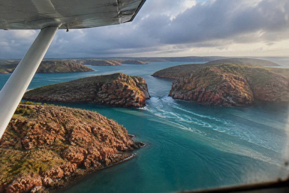 Light aircraft flying over red cliffs and turquoise sea along a remote Kimberley coastline