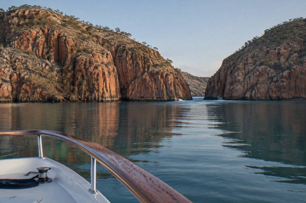 A boat crossing open water toward steep red cliff walls in a remote Kimberley bay