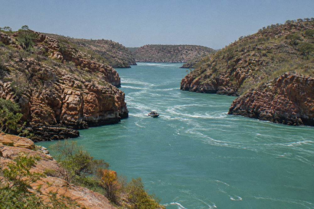 A wide tidal channel between layered red rock ranges in the Buccaneer Archipelago at midday