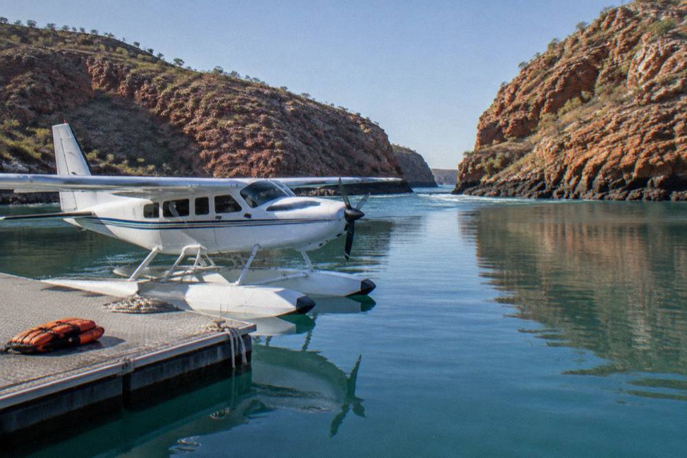 A seaplane moored at a floating pontoon beside red rock cliffs in a remote bay