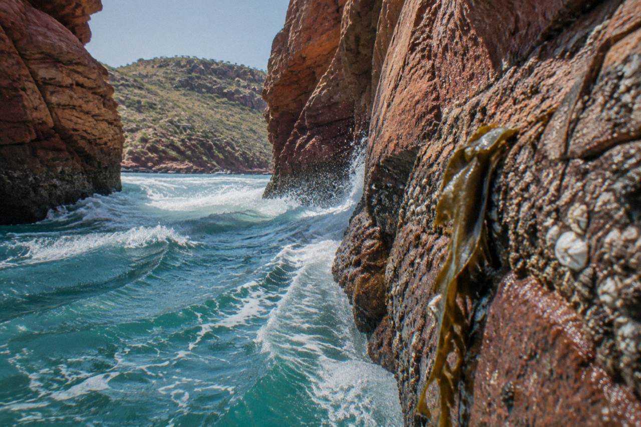 Turquoise tidal water rushing through a narrow red rock gap in a remote Kimberley bay