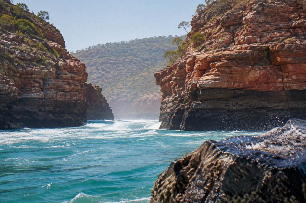 Turquoise water rushing through a narrow coastal gorge surrounded by red rock cliffs