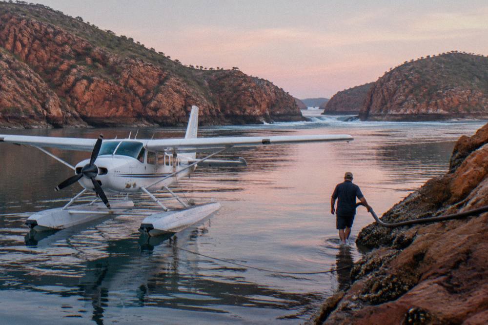 A seaplane floating on calm turquoise water beside a red rock coastline
