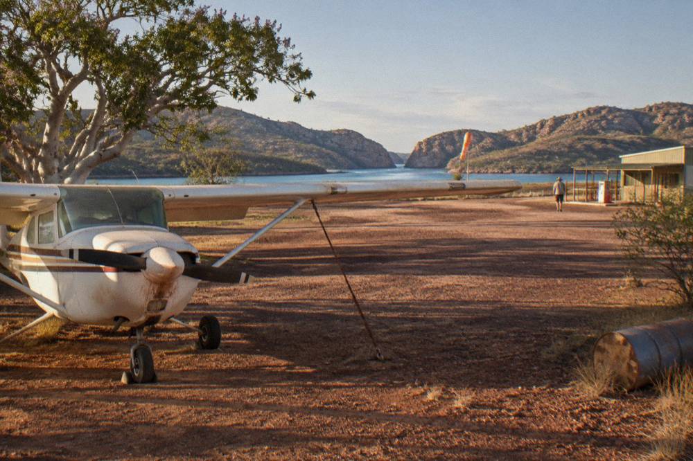 A light aircraft parked on a small regional airstrip with red earth and sparse trees