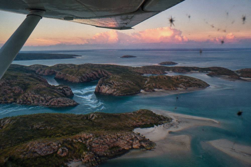 A light aircraft wing above clustered islands and pale sandbars in clear sea water
