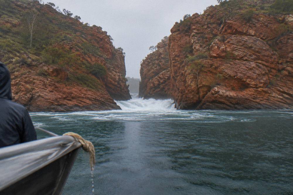 A narrow chasm of red rock with white water surging through it into a darker bay