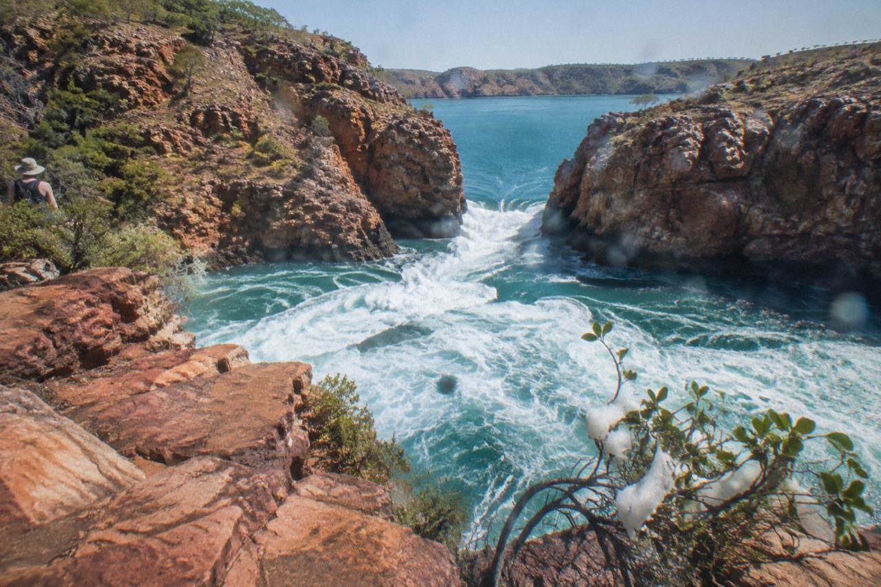 Turbulent water rushing through a narrow red rock gap in Talbot Bay under a clear sky