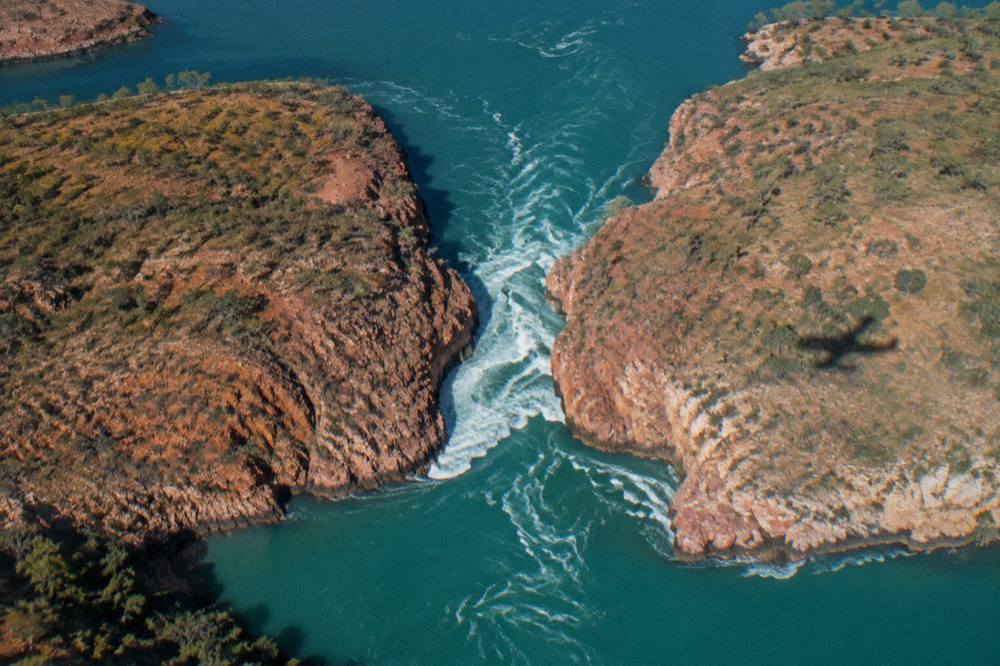 Turquoise water rushing through a narrow rocky gap between red cliffs seen from above