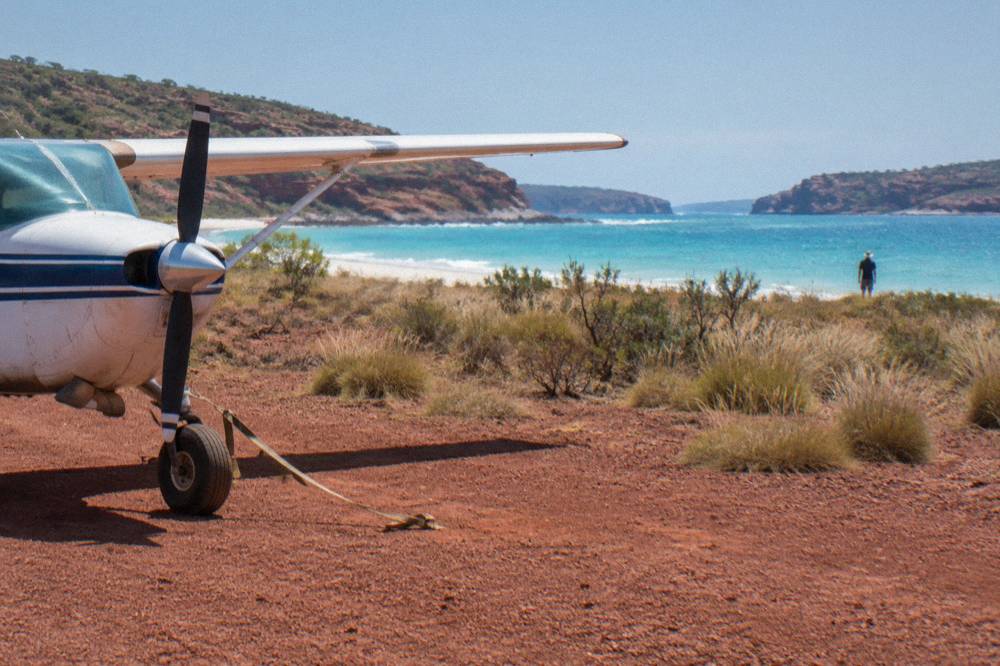 A small fixed wing aircraft parked on a red dirt airstrip near white sand and turquoise sea