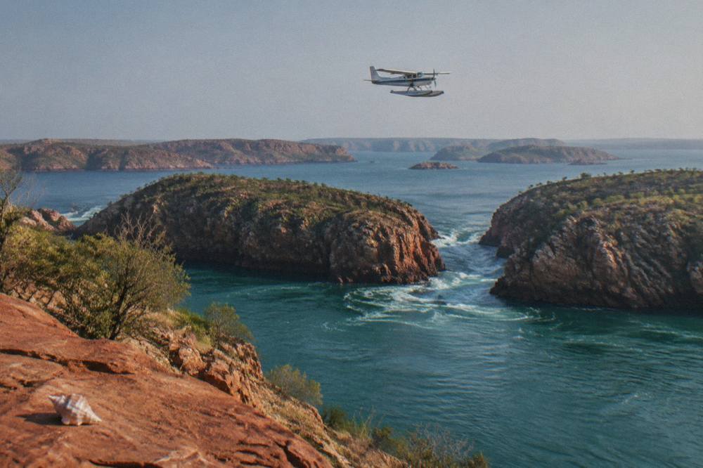A seaplane in flight over red cliffs and scattered islands along the Kimberley coast