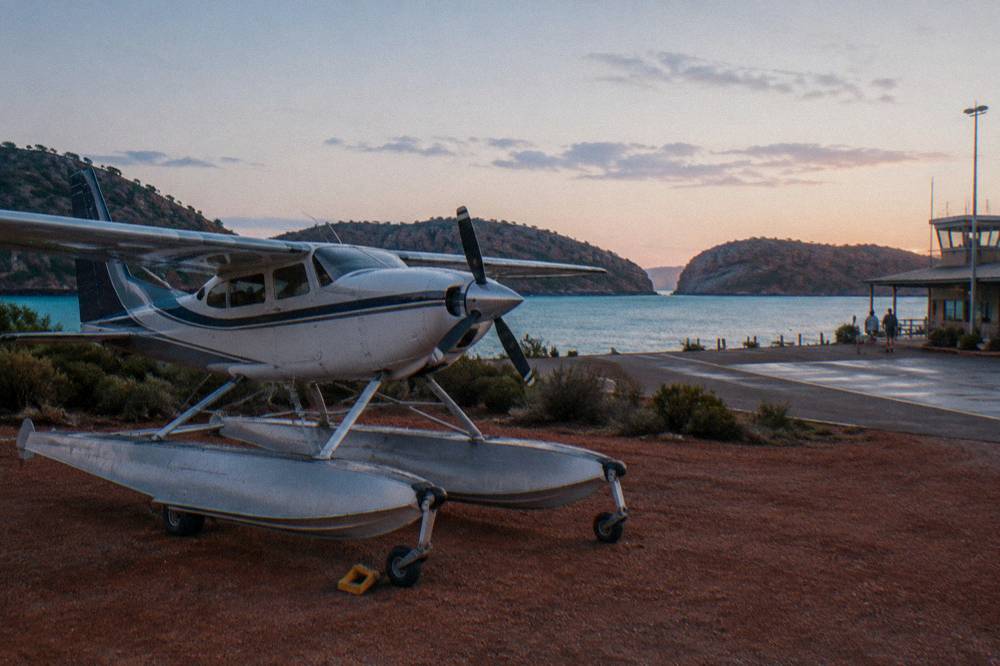 A small aircraft on a red dirt airstrip beside turquoise water at sunrise