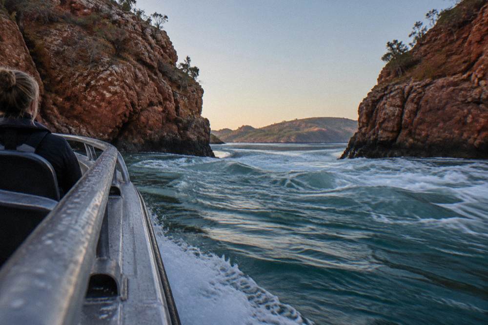 A power boat moving through a narrow gap between dark cliffs with churning white water on both sides