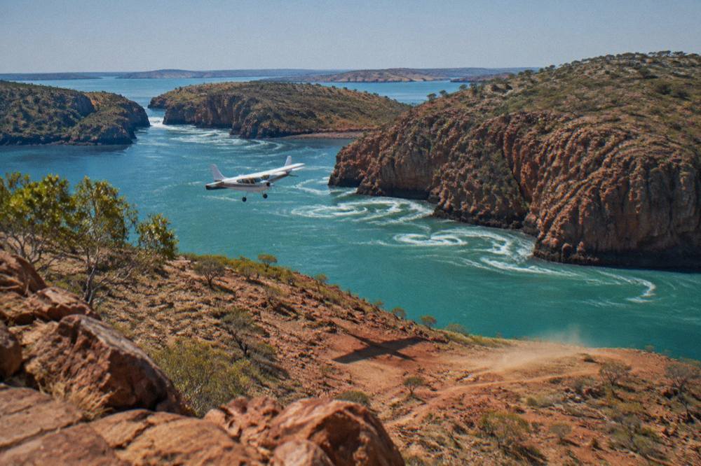 Small fixed-wing aircraft flying low over red cliffs and turquoise water in the Kimberley