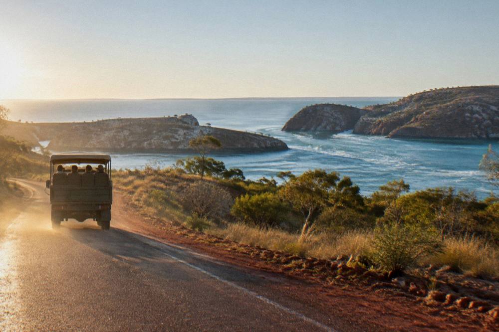 Tour vehicle on a sealed red-dirt road through pindan country toward a distant coastline