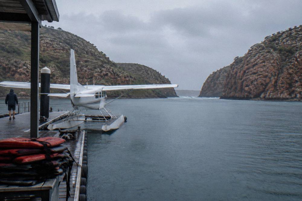 A seaplane moored beside a floating pontoon in a remote bay surrounded by red rock cliffs
