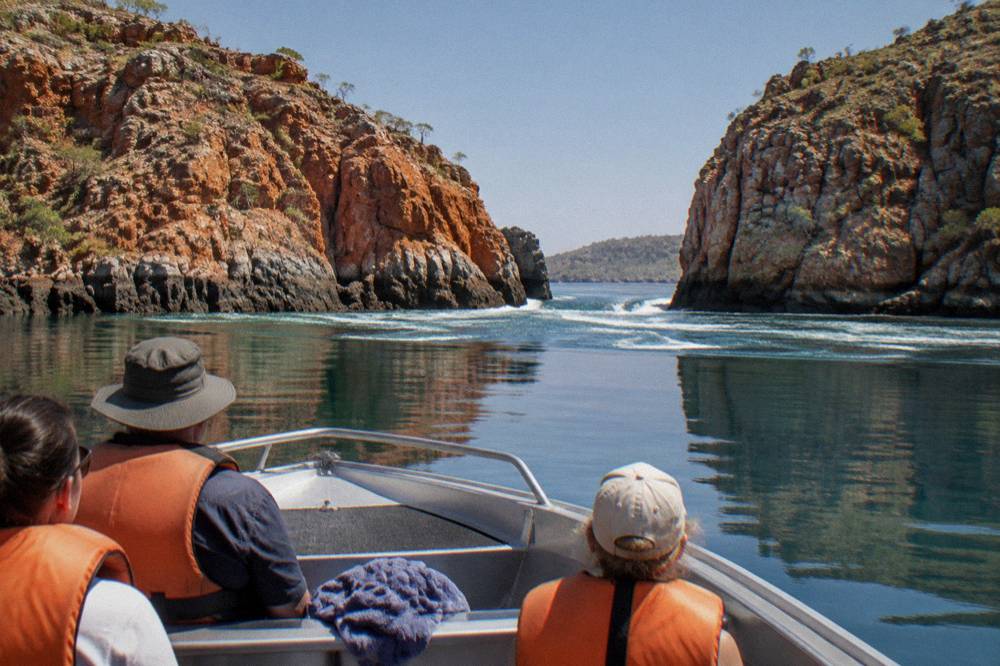 Passengers in life jackets seated in an open power boat beside red rock cliffs on calm water