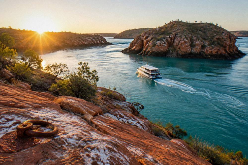 A tour boat cruising calm turquoise water between rugged red islands at sunrise in the Buccaneer Archipelago
