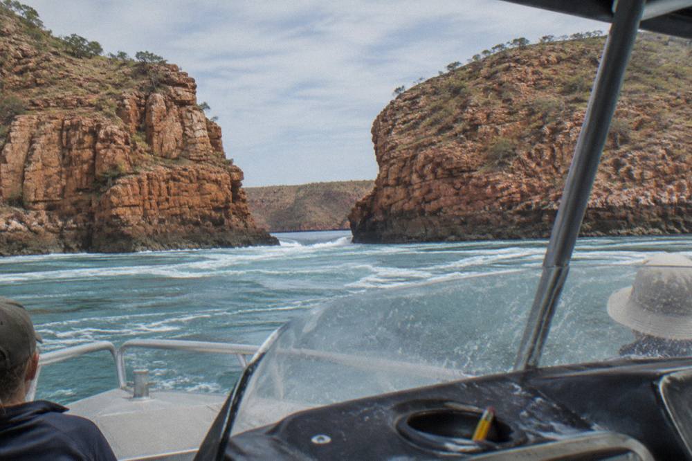 A flat-bottomed cruise boat travelling across open turquoise water toward layered red cliff walls