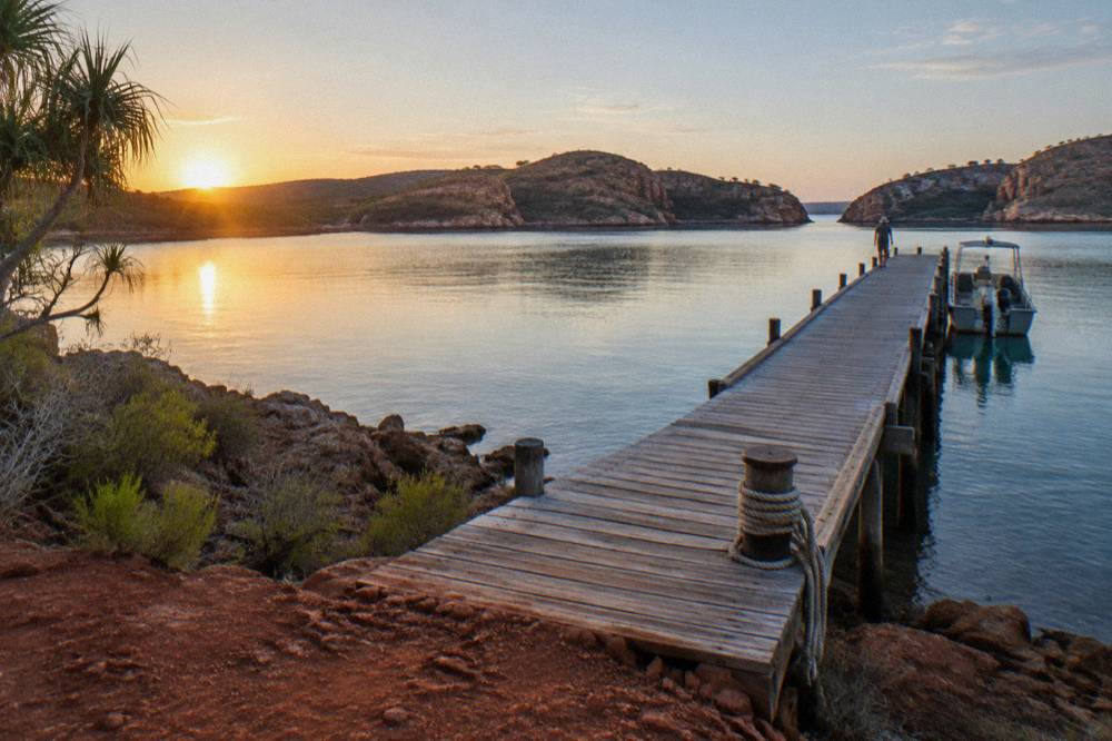 A long wooden jetty stretching out from a red dirt shoreline into calm blue water at sunrise