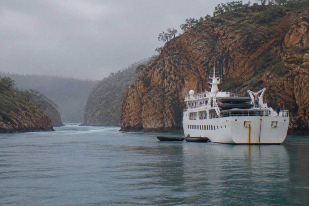 A small expedition ship anchored beside tall orange cliffs in a quiet Kimberley inlet