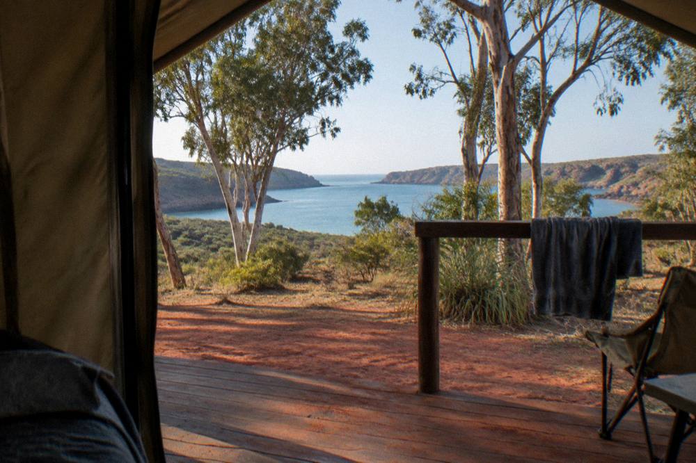 A safari tent with a shaded deck set on red dirt among scattered gum trees near a blue bay