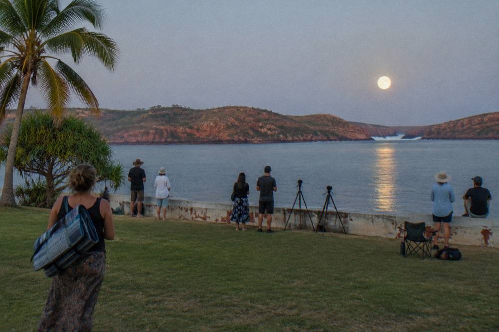 Crowd gathered at a foreshore park watching a full moon rise over calm bay waters