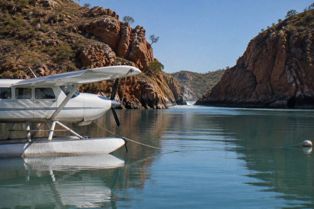 Seaplane floating on calm turquoise water beside a red rock coastline at midday