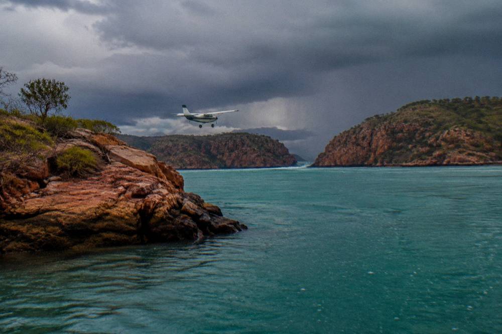 A light aircraft flying low over turquoise island-dotted water near red cliffs