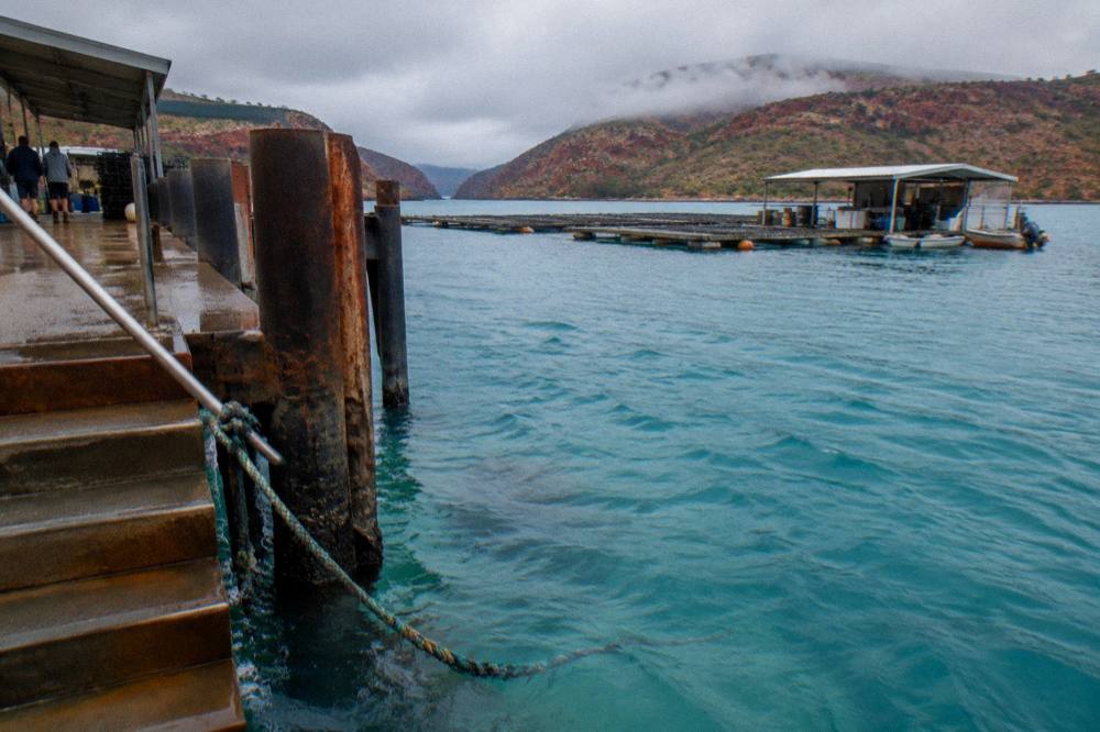 Turquoise reef water beside a pearl farm jetty with red rocky hills behind