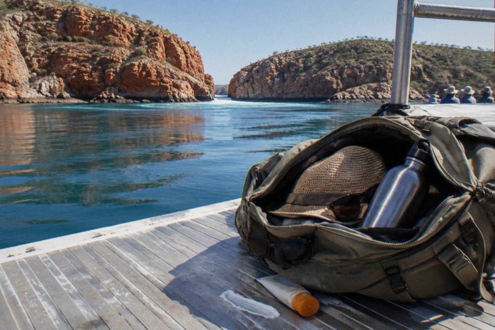 An open day bag with a sun hat, sunglasses and a water bottle resting on a wooden deck in bright tropical sunlight