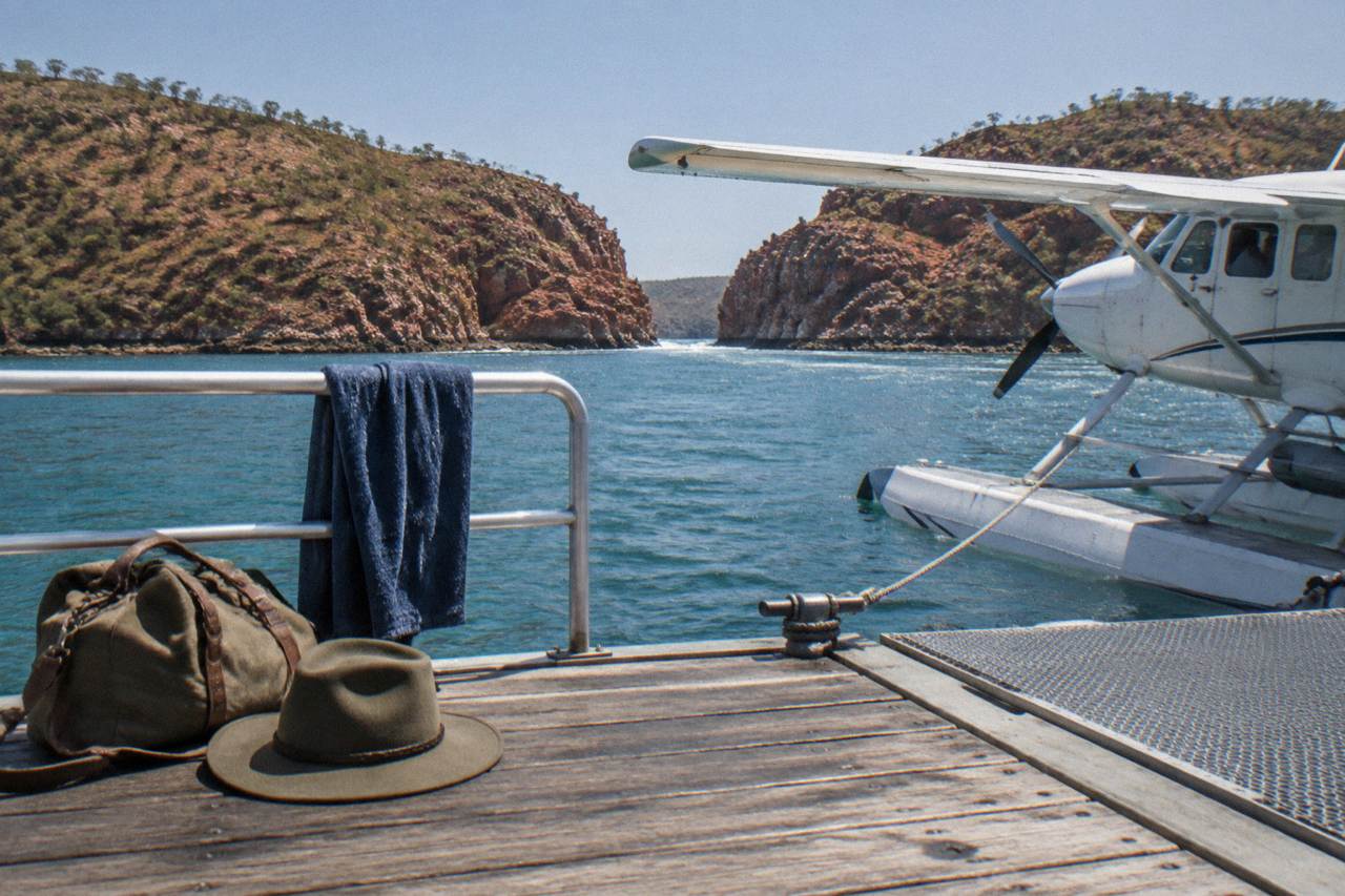 A packed day bag and hat on a wooden jetty beside a moored seaplane on turquoise Kimberley water