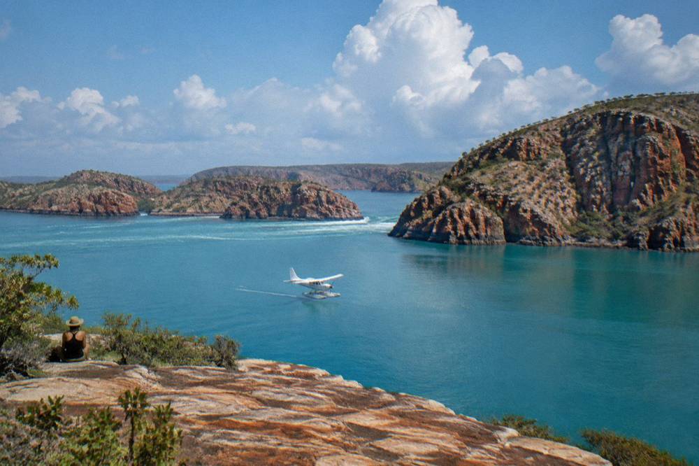 A seaplane flying low over turquoise island waters toward red cliffs in a remote archipelago